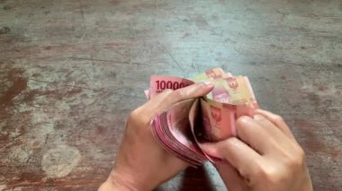 4K close-up of hands counting rupiah bills with three fingers, left hand holding stack, on rustic surface. Perfect for salary, finance, audit, savings, and banking themes.