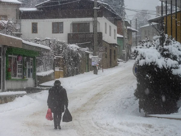 A Man Carrying Bags on Snow Covered Street in a small Bulgarian Village.