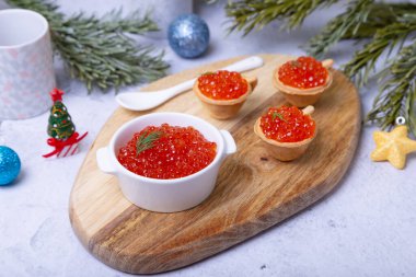 Tahta üzerinde beyaz bir kasede kırmızı havyar (somon havyarı). Arka planda havyarlı turtalar, Noel ağacı dalları, oyuncaklar ve bir mum var..