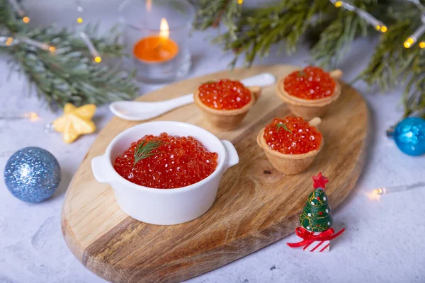 Red caviar (salmon caviar) in a white bowl on a wooden board. In the background are tartlets with caviar, Christmas tree branches, toys and a candle.