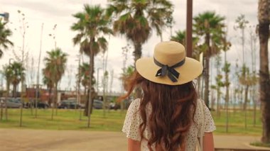 Kadın turist, şehir parkında palmiye ağaçları ve güneş ışığıyla yürümekten zevk alıyor.