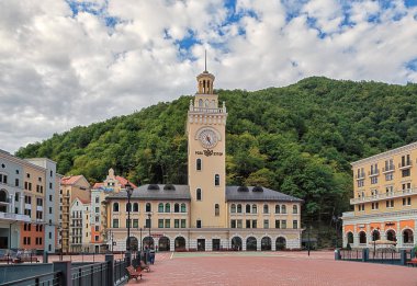 Soçi, Rusya - 8 Ağustos 2013: Bulutlar ile mavi gökyüzü altında yeşil yaz dağ ormanla çevrili Rosa Khutor dağ tatil beldesinin güzel townscape. Saat Kulesi ve ana kare doğal peyzaj