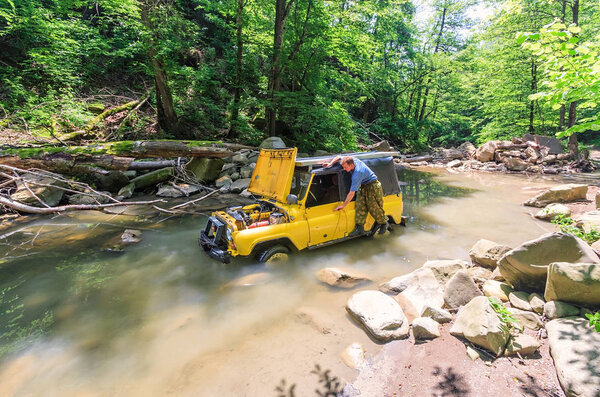 Sochi, Russia - June 9, 2009: Yellow off-road Russain car stuck in mountain river at sudden breakdown while jeeping. Driver repairs auto with lifted hood. Scenic summer forest landscape on sunny day.