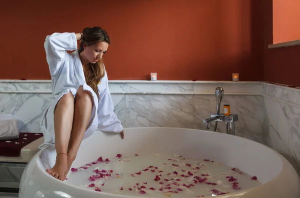 Joven hermosa mujer morena caucásica en albornoz blanco está a punto de tener bañera de hidromasaje con pétalos de rosa. Turismo de bienestar y recreación en el resort —  Fotos de Stock