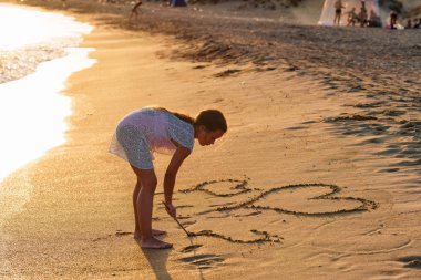 Yaz gün batımında kumlu plajda bir sopa ile kalplerin figürleri çizim küçük Kafkas kız