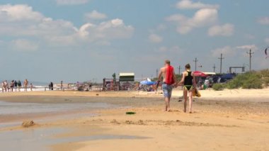 Anapa, Rusya - 11 Haziran 2017: Bugaz'daki kumlu Karadeniz sahilinde el ele tutuşan ve bira şişeleri tutan genç erkek ve kadın çift. Güneşli yaz gününde kumlu deniz kıyısında kırma sörf dalgaları. 4k görüntü