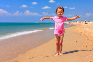 Mavi gökyüzü ve deniz sörfü arka plan güneşli bir günde yaz sahil kumlu plajda duran sevimli küçük Kafkas kız, gülümseyerek ve el sallayarak. Anapa resort, Rusya Federasyonu.