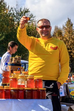 Gülümseyen adam, beyaz dağ kırsalında açık hava pazarında güneşli bir günde içmeye hazır bir bardak mead tutar ve bal satışa çıkar