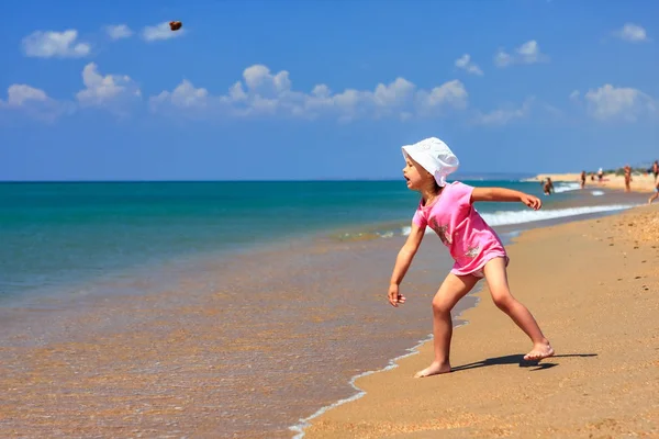 Cute little active Caucasian girl on sandy beach at summer seaside throwing stone into the sea on sunny day on blue sky background. Anapa resort. Russia. — Stock Photo, Image