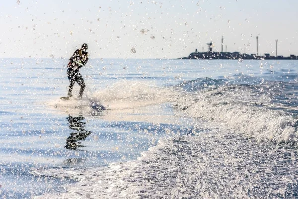 Wakeboarder se desliza sobre la superficie del mar remolcado por un barco a motor con salpicaduras que se dispersan al atardecer. El wakeboarding como deporte extremo al aire libre y actividad de ocio en el resort de verano. Utrish, Anapa, Rusia . — Foto de Stock