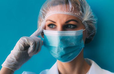 Tıbbi şapkalı, maskeli, beyaz önlüklü ve mavi arka planda eldivenli genç kadın portresi. Şakakta parmağı olan bir doktor, koronavirüs salgınında yarını düşünüyor.