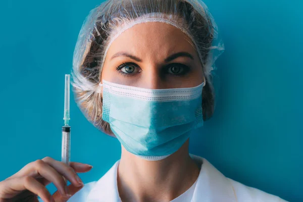 Tıbbi başlık ve maske takmış, elinde beyaz önlük ve şırıngayla mavi arka planda genç bir kadın portresi. Doktor, Coronavirüs salgınında zor bir günde iğne yaptıktan sonra yorgun düştü.
