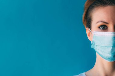 Mavi arka planda, tıbbi maskeli genç bir kadının yakın plan yarım yüzü. Koronavirüsten koruma. Cerrahi maskeli kız. Klinikteki kadın doktor. Dönem salgınında evde kalma kavramı.