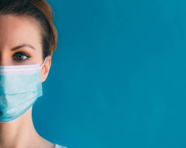 Mavi arka planda, tıbbi maskeli genç bir kadının yakın plan yarım yüzü. Koronavirüsten koruma. Cerrahi maskeli kız. Klinikteki kadın doktor. Dönem salgınında evde kalma kavramı.