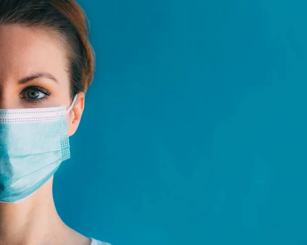 Mavi arka planda, tıbbi maskeli genç bir kadının yakın plan yarım yüzü. Koronavirüsten koruma. Cerrahi maskeli kız. Klinikteki kadın doktor. Dönem salgınında evde kalma kavramı.