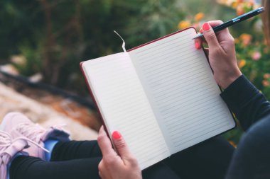 Kadın ellerindeki boş defterin yanında bir kalemle parkta otururken çekilmiş fotoğrafı. Kadının elleri boş bir defter tutuyor. Bütün rüyalarını bir deftere yaz. Kalemle el yazısı. Uzay metni