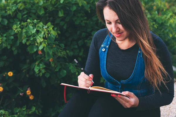 Young woman on bench in park with notebook and pen on green background. Wish Marathon. Writing wishes and dreams in a notebook. Life with a clean slate. Planning a new week. Life after quarantine