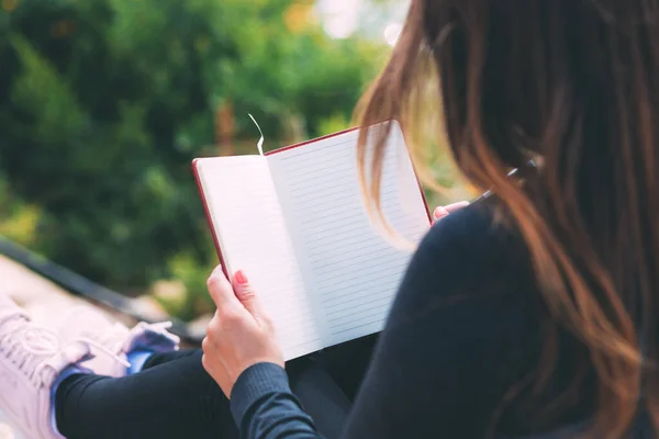 Side photo of blank notebook in female hands together with a pen, sitting in a park. Woman\'s hands are holding blank notebook. Write all your dreams in a notebook. Hand writing with pen. Space text