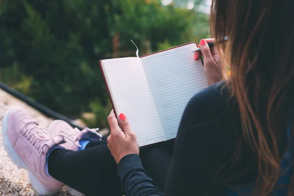 Side photo of blank notebook in female hands together with a pen, sitting in a park. Woman\'s hands are holding blank notebook. Write all your dreams in a notebook. Hand writing with pen. Space text