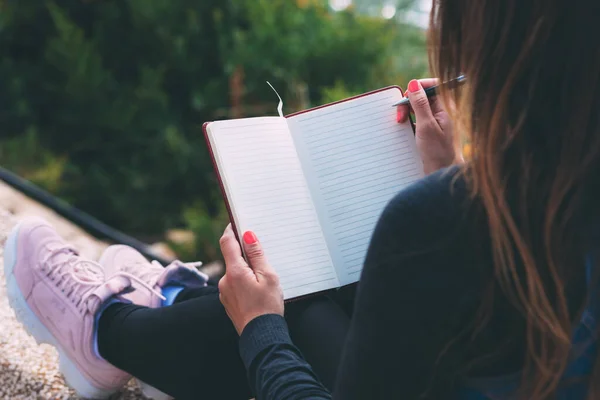 Kadın ellerindeki boş defterin yanında bir kalemle parkta otururken çekilmiş fotoğrafı. Kadının elleri boş bir defter tutuyor. Bütün rüyalarını bir deftere yaz. Kalemle el yazısı. Uzay metni