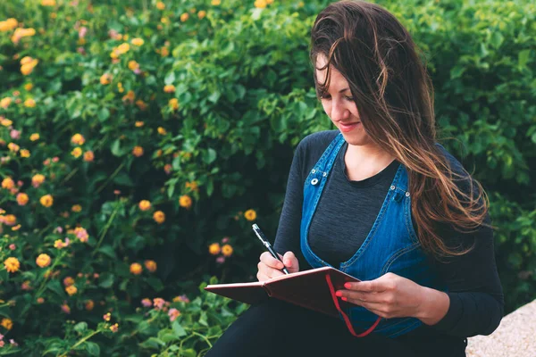 Parktaki bankta yeşil arka planda defter ve kalemle gülümseyen genç bir kadın. Dilek Maratonu. Not defterine rüya yazmak. Temiz bir sayfa açarak. Yeni bir hafta planlıyorum. Karantina sonrası yaşam