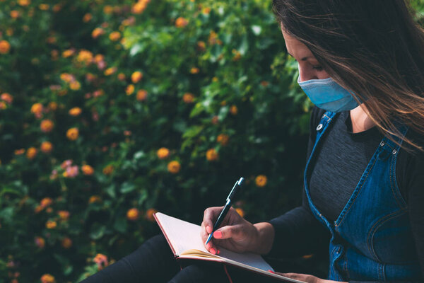 A young woman in a medical mask sits in a park and writes something in a notebook. Life after quarantine during the coronavirus epidemic pandemic. New reality. Respiratory protection. Green background