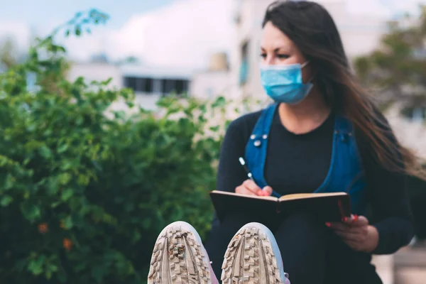 Not defteri ve kalemle parkta maske takmış bulanık genç bir kadın yazmış. Ön planda bulanık portre ve spor ayakkabılar. Koronavirüs enfeksiyonu sırasında karantina sonrası yaşam. Yeni gerçeklik