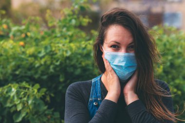 Yüzünde koruyucu tıbbi maske takan, yüzünün yakınında el ele tutuşan açık havada şaşırmış bir kadının yakın plan portresi. Cerrahi maskeli güzel esmer. Karantina sonrası yaşam, koronavirüs salgını