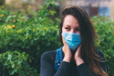Yüzünde koruyucu tıbbi maske takan, yüzünün yakınında el ele tutuşan açık havada şaşırmış bir kadının yakın plan portresi. Cerrahi maskeli güzel esmer. Karantina sonrası yaşam, koronavirüs salgını