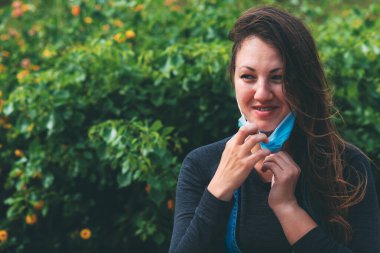 Genç bir kadın gülümser ve mavi bir tıbbi maske takar ve karantinanın sonunda, parkta, dışarıda sevinir. Koronavirüs salgını sırasında karantina sonrası yaşam. Yeni gerçeklik. Yeşil arkaplan