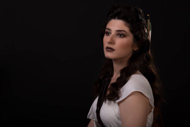 Siyah arka planda beyaz elbiseli okçu bir kadının yakın plan portresi. Yunan tanrıçası Artemis. Stüdyo fotoğraf çekimi. Uzun siyah saçlı bir esmer için profesyonel makyaj ve saç stili. Genç kadın.