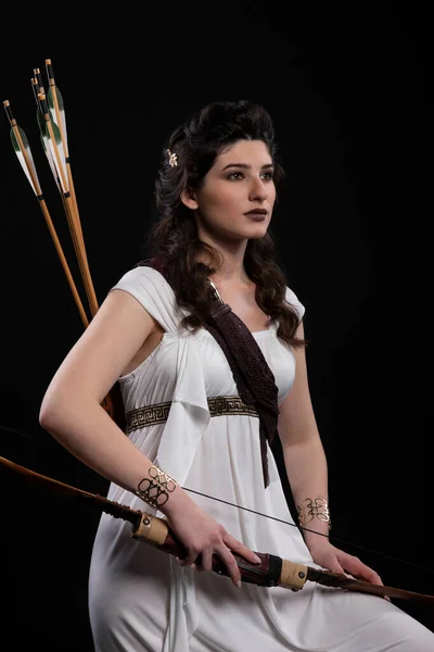 Siyah arka planda ok ve yay olan beyaz elbiseli bir okçu kadın portresi. Yunan tanrıçası Artemis. Stüdyo fotoğraf çekimi. Uzun siyah saçlı bir esmer için profesyonel makyaj ve saç stili.