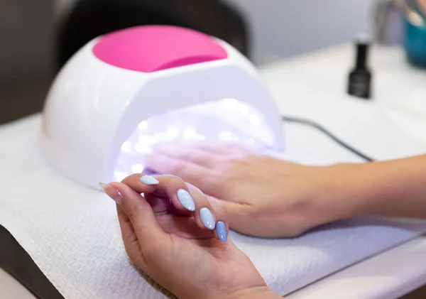 Close-up beautiful woman hands and lamp for nails on table. Nail gel salon. UV lamp. Process of drying gel on nails. Beauty and self care. Modern technology in nail salon. Nail care after quarantine