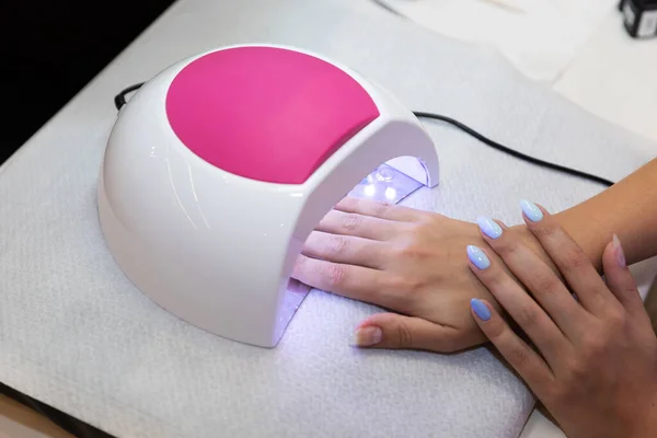 Close-up beautiful woman hands and lamp for nails on table. Nail gel salon. UV lamp. Process of drying gel on nails. Beauty and self care. Modern technology in nail salon. Nail care after quarantine