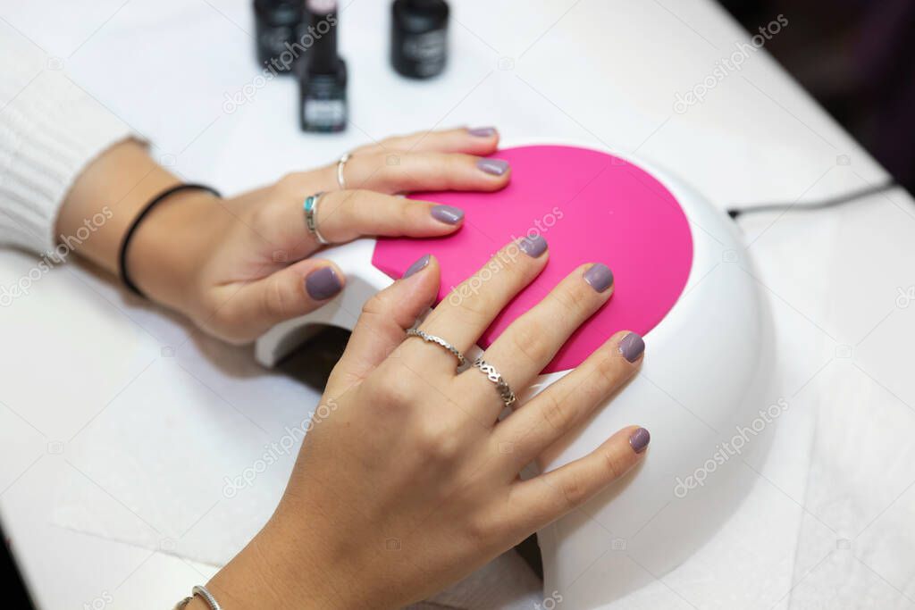 Ready manicure in purple. Hands of a young girl with a gentle purple manicure on a machine for ultraviolet drying gel varnish  in the salon. Nail care after quarantine.