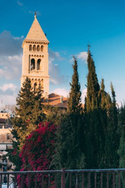 Kudüs Eski Şehir, Hristiyan Mahallesi. Kudüs 'ün çatılarından Hristiyan kilisesinin manzarası. Kudüs Günü. Eski Kudüs Şehri. Yeşil ağaçlar ve mor bougainvillea çalıları. Mavi gökyüzü, bulutlar