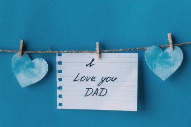 Babalar Günü. Siyah el yazısıyla yazılmış tebrik kartı içeren beyaz defter. Babanı seviyorum. Mavi arka planda izole edilmiş çamaşır mandallarıyla asılı duruyor.