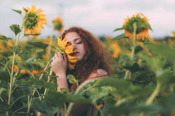 Beautiful young girl with red wavy hair and freckles in stripped colourful dress enjoying nature on the field of sunflowers. The concept of summer and sun. Sunflower season. Pressed against sunflower