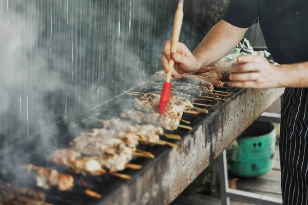 Adamın bulanık elleri, uzun dikdörtgen barbeküde soslu fırçayla, açık havada dumanla eti yağlıyor. Yaz barbeküsü pişirme. Çiğ sebzeli barbekü. Terasta barbekü.