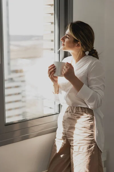 Pencerenin yanında kahve ya da çay içen genç ve güzel bir kadın. Hayal kur ve rahatla. İyi günler. Sabah kadın elinde bir kupayla güneşin doğuşunu seyrediyor ve mükemmel bir sabah geçiriyor.