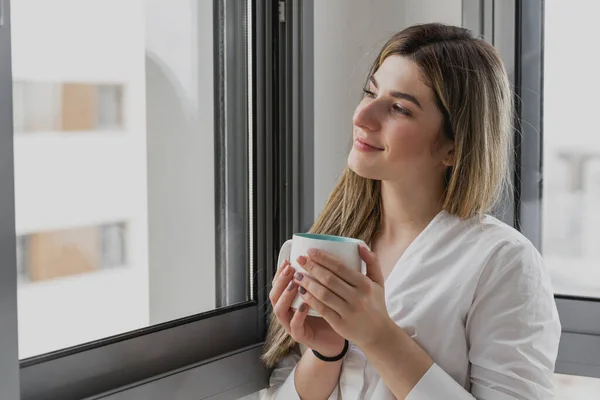 Young beautiful woman near window with cup of coffee or tea at home. Dream and relax. Good day. Woman in the morning is holding a mug and looking at the sunrise and having a perfect cozy morning