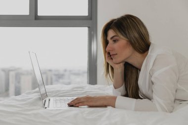 Yatak odasındaki yatağın üzerinde dizüstü bilgisayar kullanarak rahatlayan genç bir kadın. Çevrimiçi sohbet. Evden çalışıyorum. Sosyal uygulamaları kontrol eden bir kadın. İnternetten yemek sipariş ettim. Koronavirüs salgını döneminde karantina