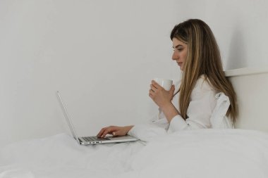 Genç ve güzel bir kadın yatak odasındaki yatağın üzerinde dizüstü bilgisayar kullanarak çay ya da kahveyle rahatlıyor. Evden çalışıyorum. Sosyal uygulamaları kontrol eden bir kadın. Çevrimiçi sipariş yemekleri