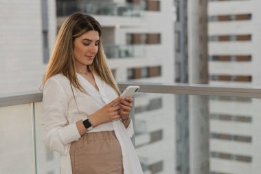 Balkonda cep telefonu kullanan genç bir kız. İş kadınları balkonda durup akıllı telefon kullanıyorlar. Beyaz elbiseli olumlu bir kadın sosyal uygulamalara bakıyor.