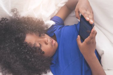 Küçük Afrikalı çocuk yatakta akıllı telefonuyla oynuyor.