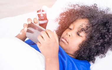 Küçük çocuk cep telefonuyla çalar saatle yatakta oynuyor.