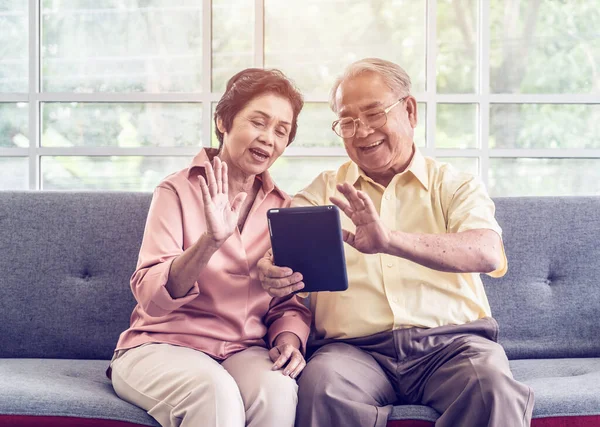 Senior couple is waving to family on Video Call communication technology on tablet on a sofa at home