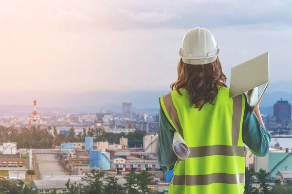 Female Civil engineer with computer and blueprint is standing infront Japan industry city in background.