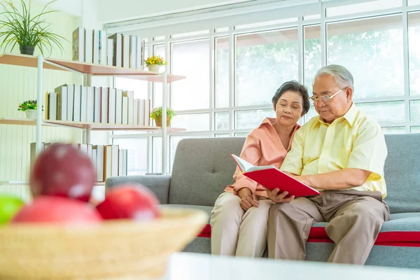 Happy retired asian couple reading book on a couch in living room with fresh fruit on the table to healthy eating and lifestyle concept.