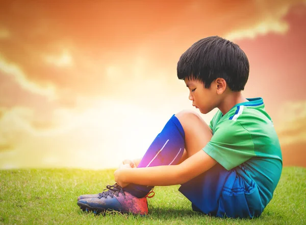 Sad soccer kid sitting on a field godlen sky for disappointment in Football sport playing.